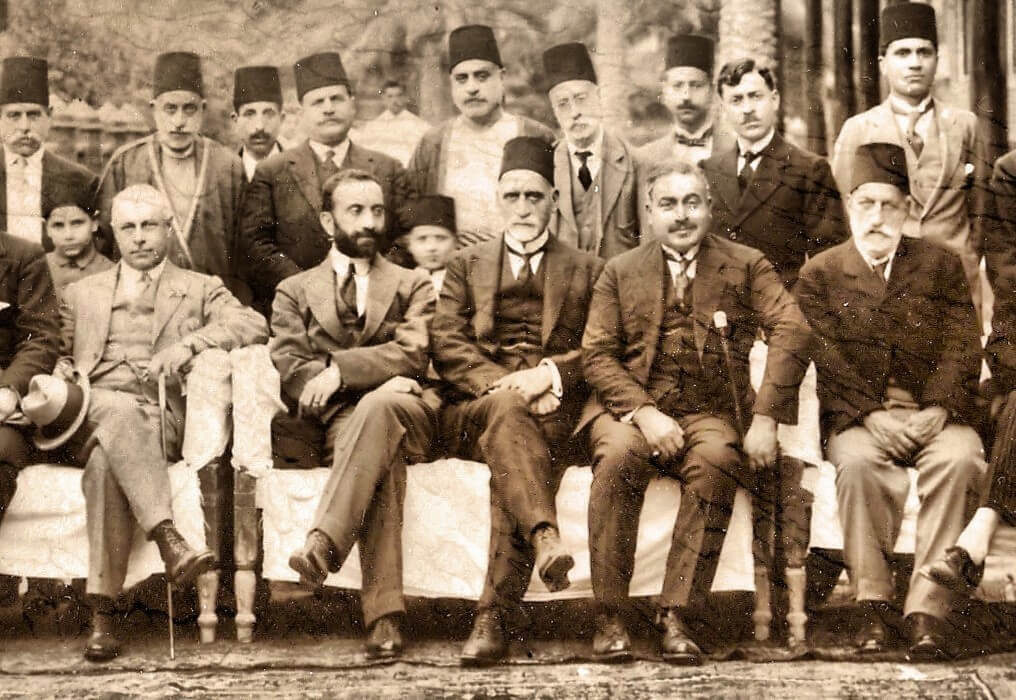 A black and white photo from the 1920s depicting Sir Sassoon Eskell, first Minister of Finance of the Hashemite Kingdom of Iraq, is seated in the center, alongside King Faisal I immediately to his left. The tycoon, Senator Menahem Saleh Daniel, is seated on the far right.